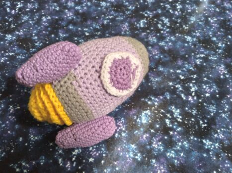 A crocheted rocket in purple, gray and white with yellow flames on the bottom against a starry sky